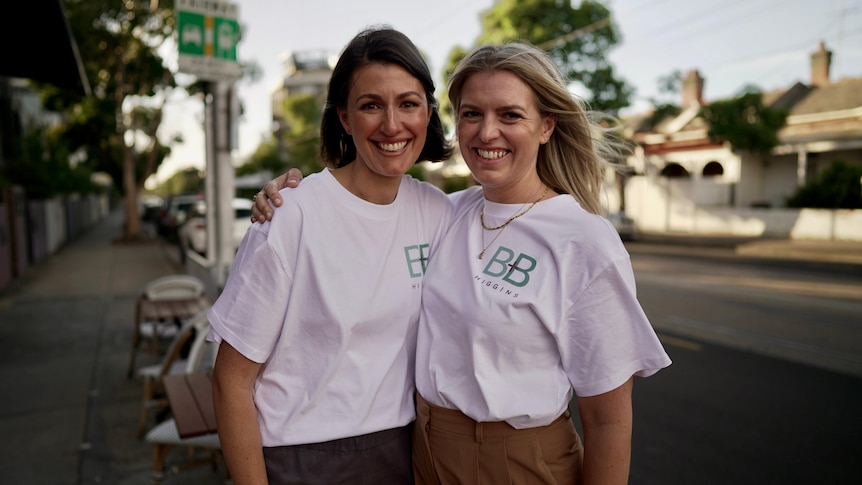 Two white women wearing white political campaign t shirts stand and smile by the side of a road