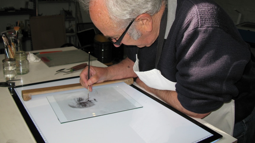 Photograph of an older man painting a face on glass, with some art supplies around him. Profile shot, gray hair, balding