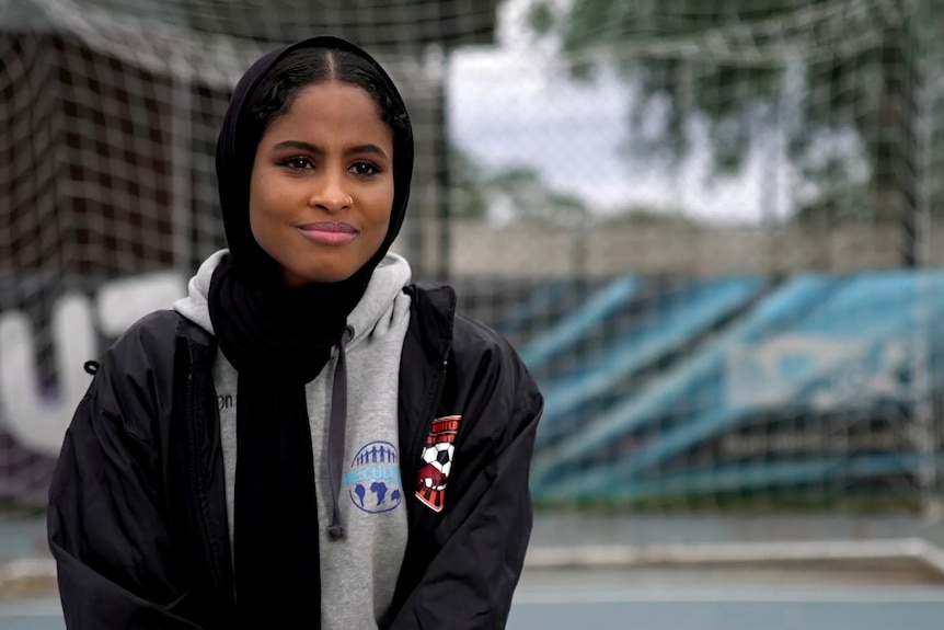 A young woman wearing a dark head scarf, a grey hoodie and a dark football jacket smiles while looking into the distance 