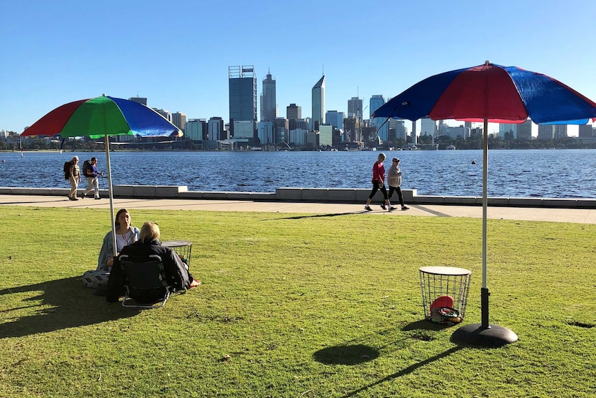 People sitting on grass on the South Perth foreshore, while others walk along the riverside.