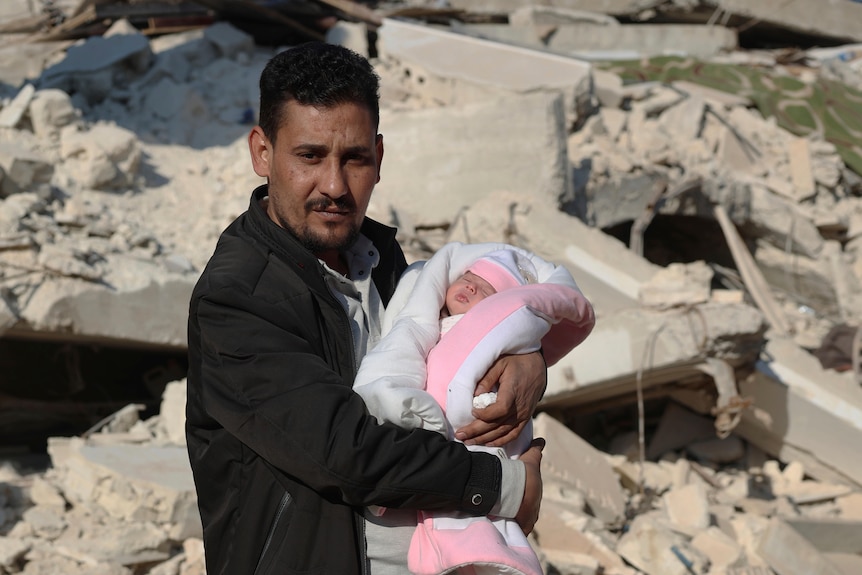 A man stands in front of a pile or rubble while holding a baby