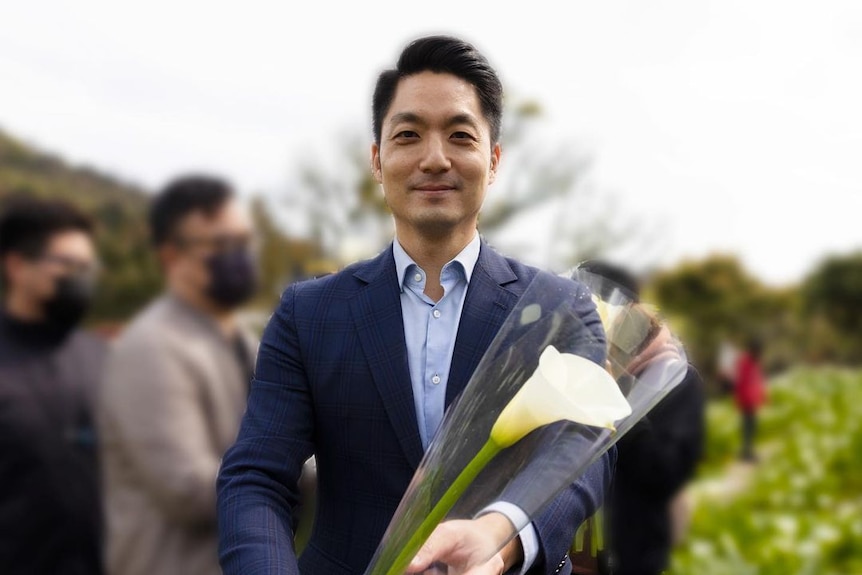 A handsome young man in a suit holding a white lily to the camera 