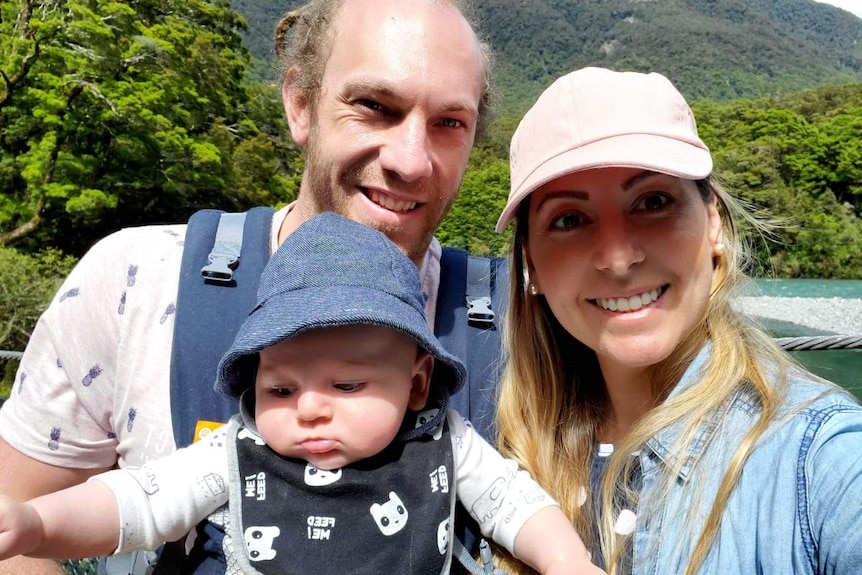 Travis wears a baby harness holding Oliver, while Brenda stands next to them.