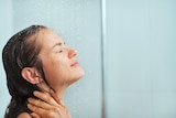 A close-up of a woman with long, brown hair with her eyes closed, standing underneath a shower head.
