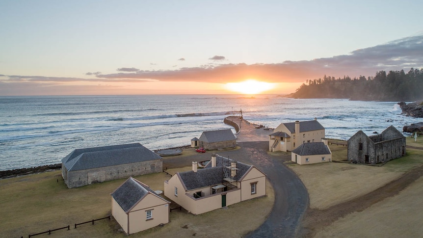 The sun sets over the water off Norfolk Island. On the land in the foreground, historic buildings face the ocean.