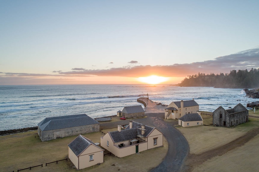 The sun sets over the water off Norfolk Island. On the land in the foreground, historic buildings face the ocean.