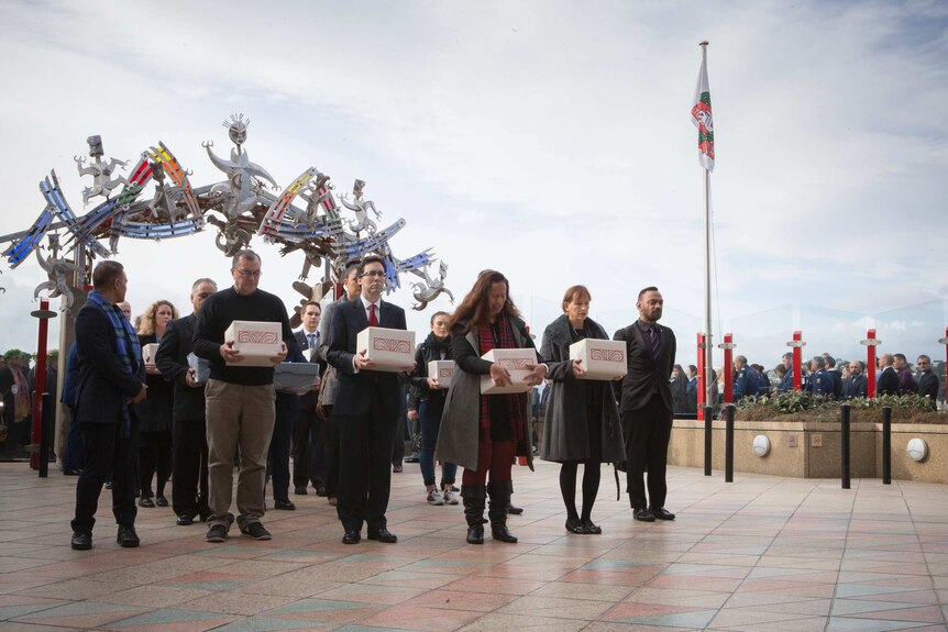 A group of solemn people holding boxes containing the remains of ancestors during a repatriation ceremony.