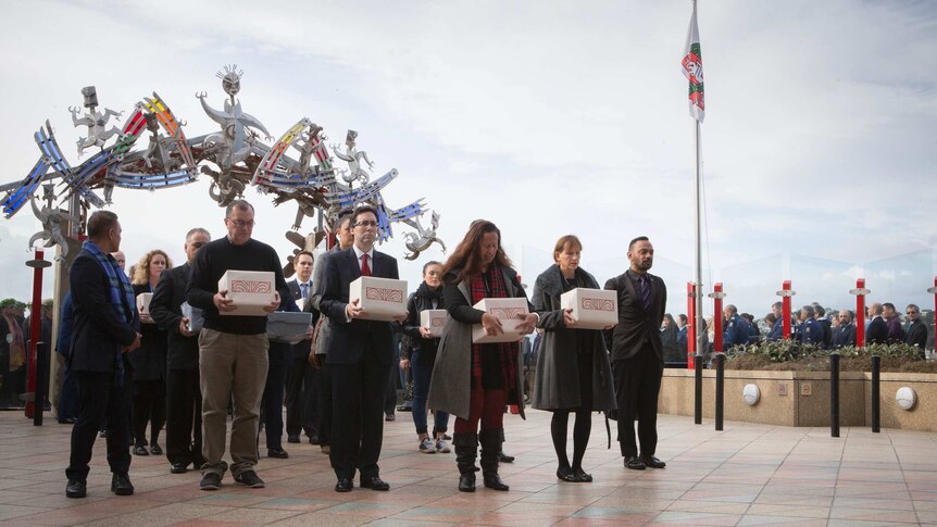 A group of solemn people holding boxes containing the remains of ancestors during a repatriation ceremony.