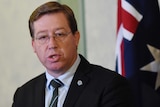 Head shot of a middle-aged man, wears shirt, dark jacket, black striped tie, glasses, speaks in front of Australian flag.