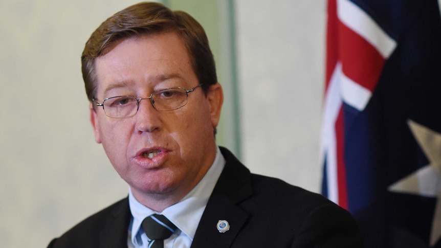 Head shot of a middle-aged man, wears shirt, dark jacket, black striped tie, glasses, speaks in front of Australian flag.