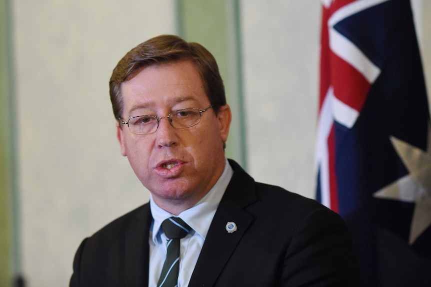 A man in dark suit and tie stands in front of the Australian flag and speaks.