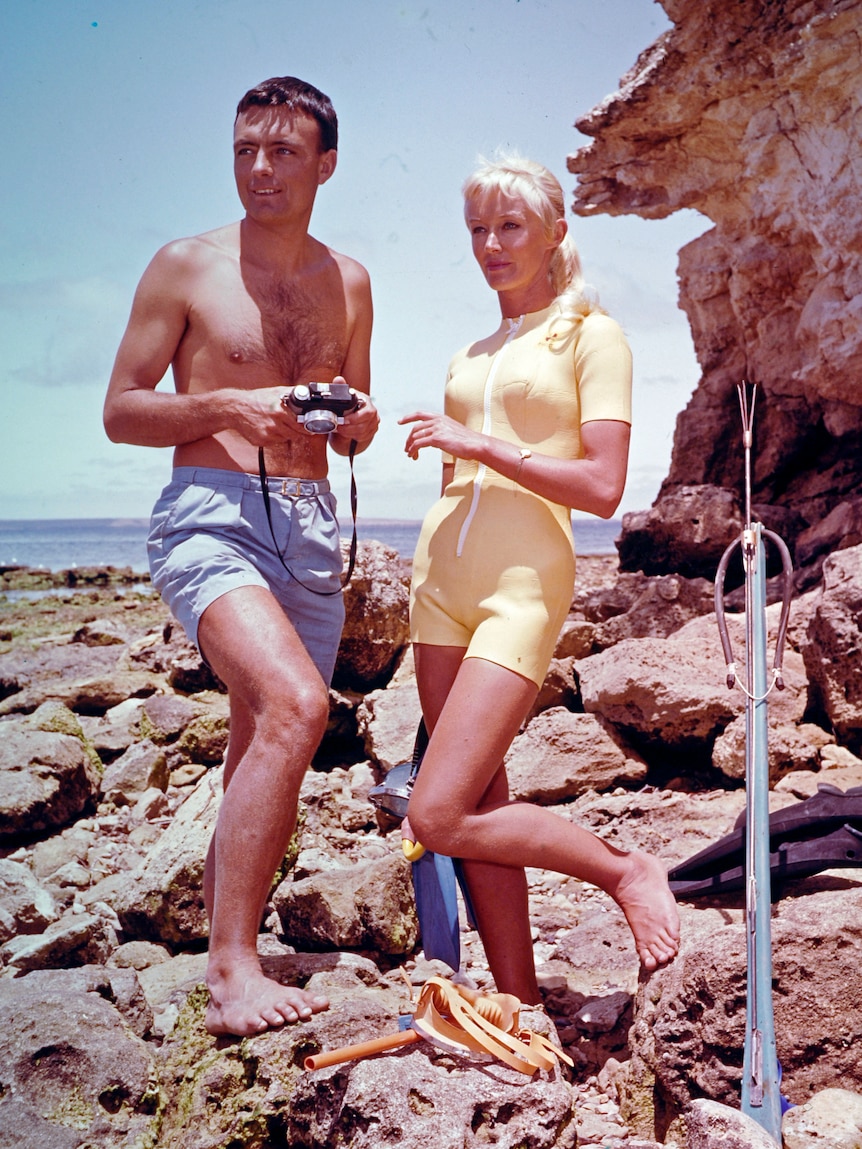 Man in swimmer trunks and woman in a yellow swimsuit stand on a boat besides rocks