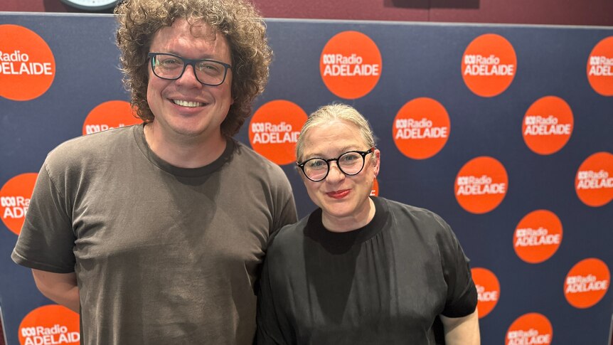man in black t-shirt with curly hair and spectacles standing nest to woman in black t-shirt with spectacles and grey hair 
