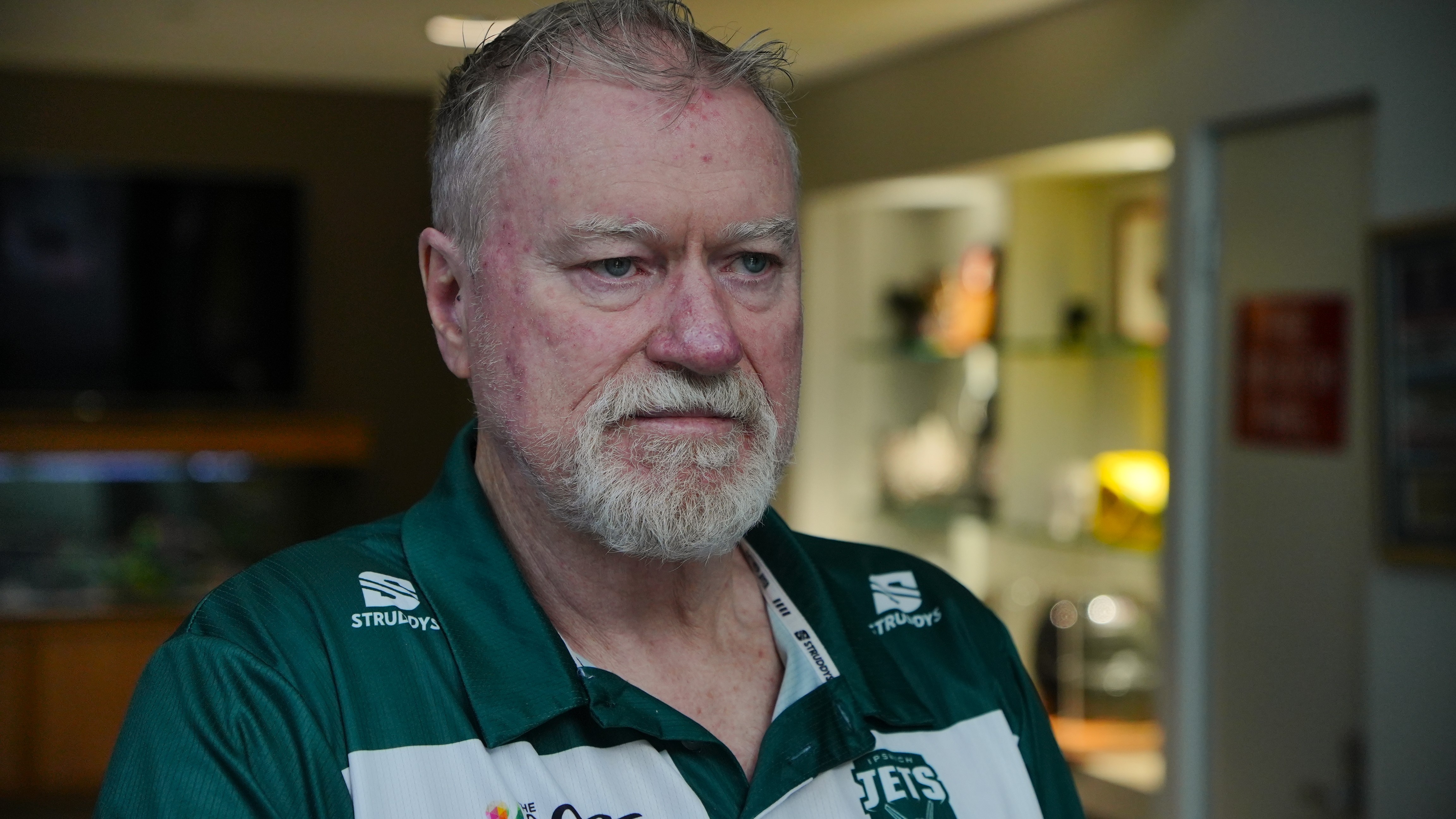 A man in a dark green polo shirt looks off camera. He has thinning grey hair and sun damage on his nose.