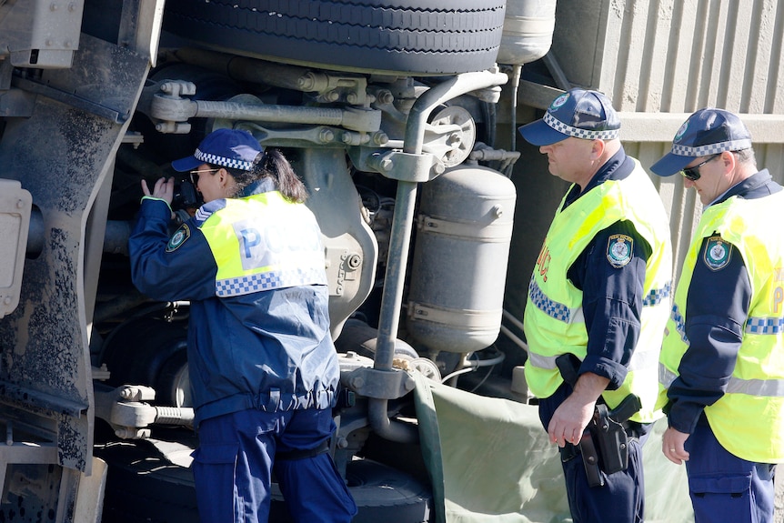 Three police officers examine the underside of a bus that has turned on its side.