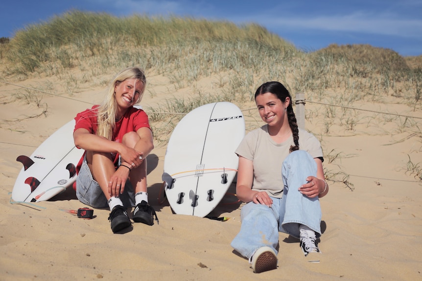 Two teenage girls sit with their surfboards in front of a sand dune.