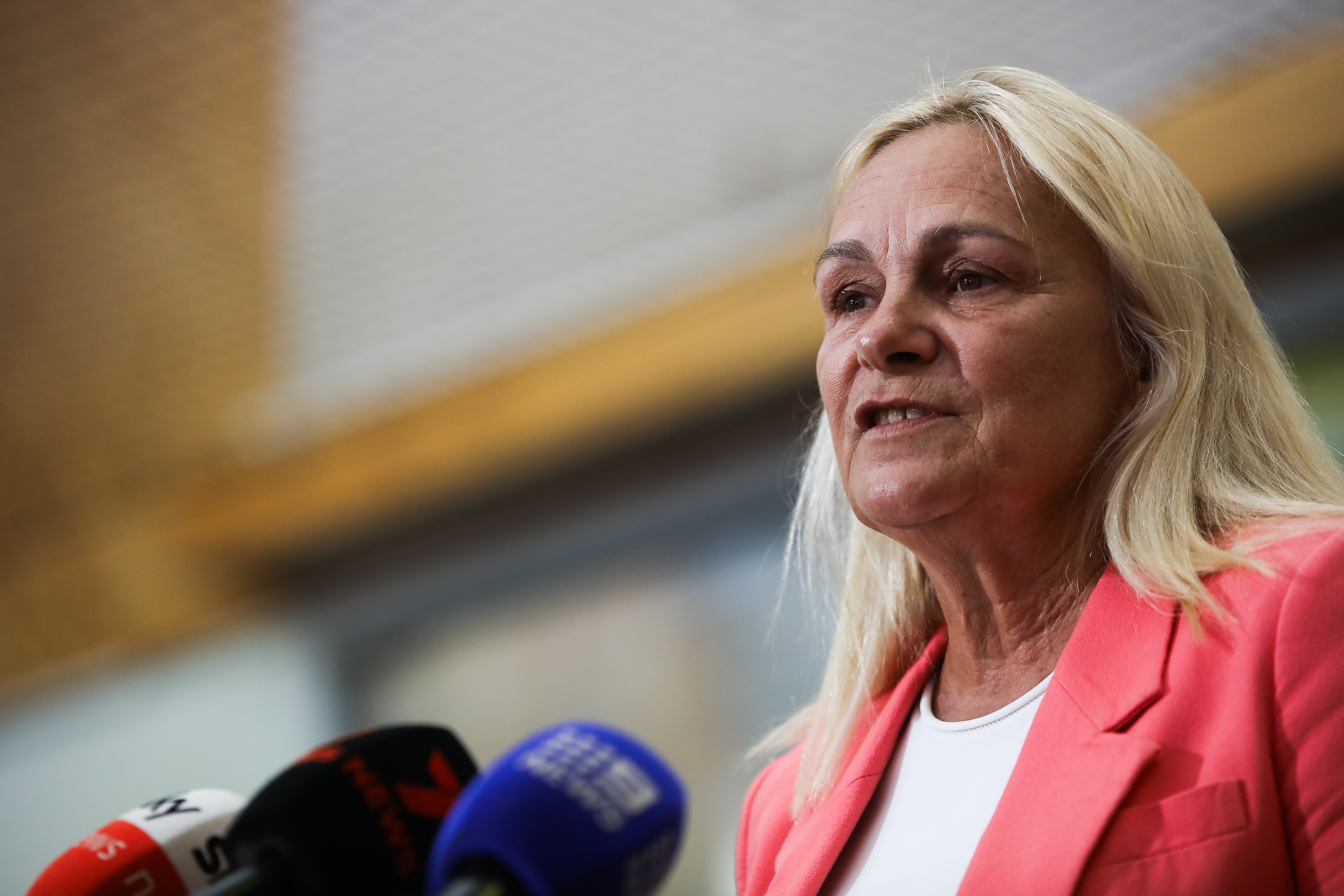 Una vista lateral en primer plano de Sabine Winton hablando ante micrófonos en una conferencia de prensa. Lleva una chaqueta rosa y un top blanco.