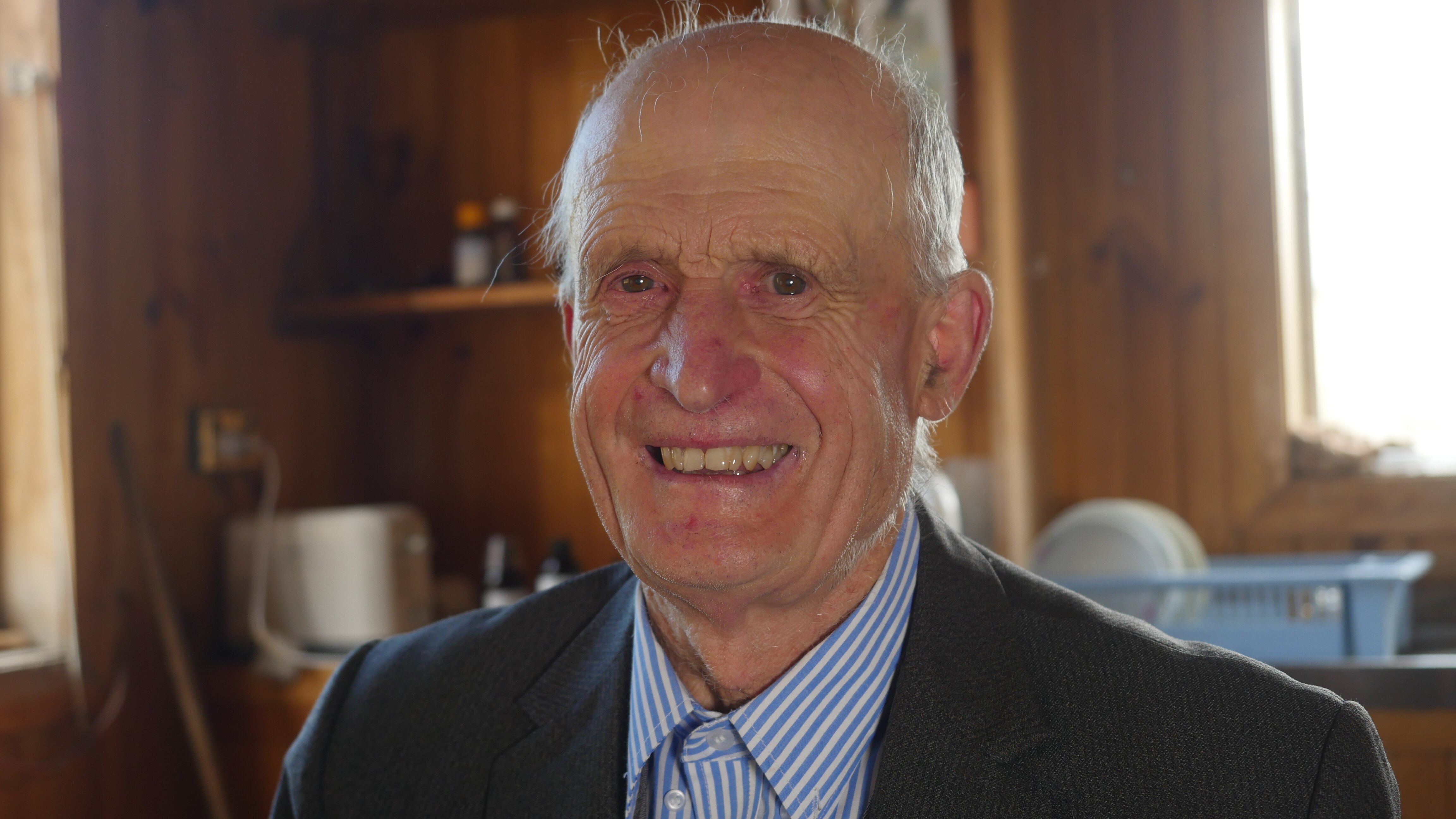 man in suit smiles at camera from his pine clad kitchen 