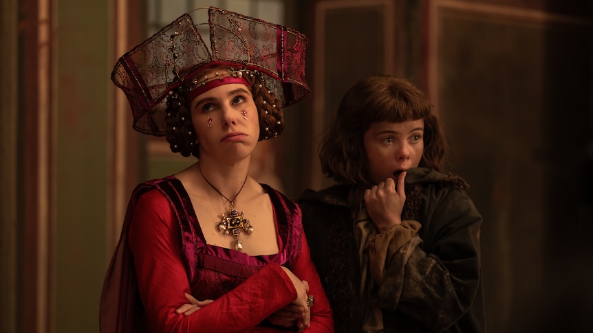 Zosia, left, wears a deep red dress and headdress and looks to the left, arms crossed and disgruntled, with Saoirse right.