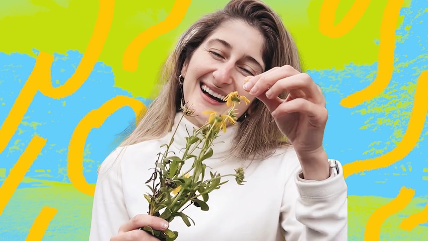 A young woman with long blonde-brown hair in a white jumper, smiles and laughs while plucking a petal off a yellow flower