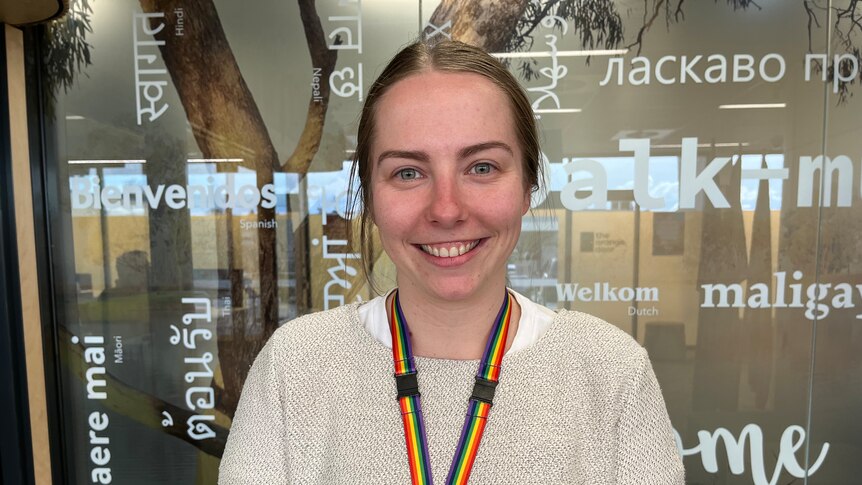 A young woman with tied-back brown hair, wearing a rainbow lanyard, smiles