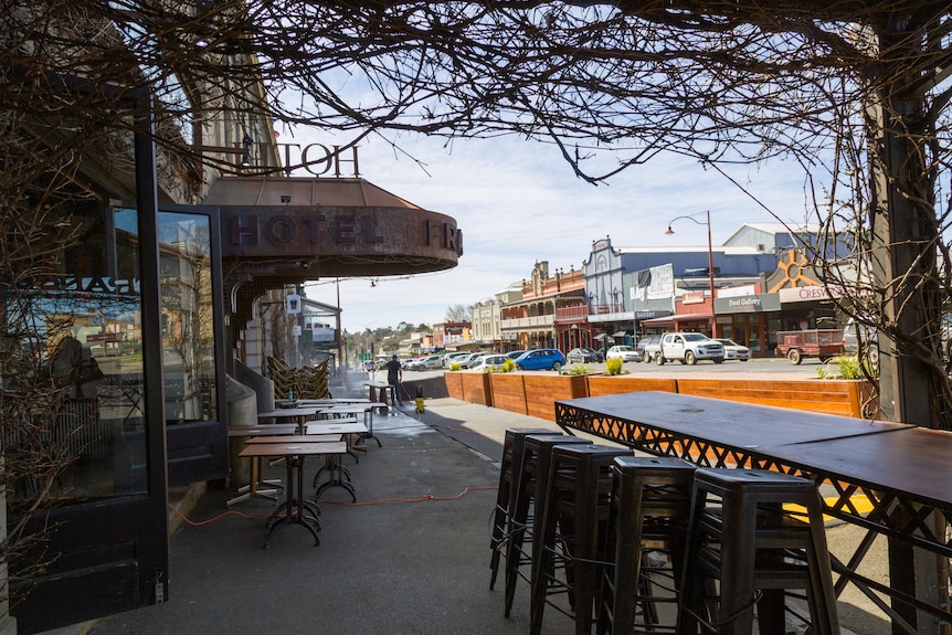 Outdoor tables, chairs placed under a vine, others under a marquee labelled hotel, with the sunny town in foreground.