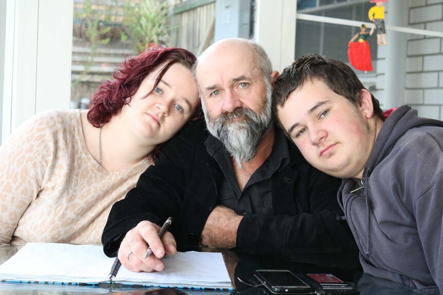 Len Twigg sits at a table with a pen in hand and and open book, next to his children Jess and Michael Twigg.