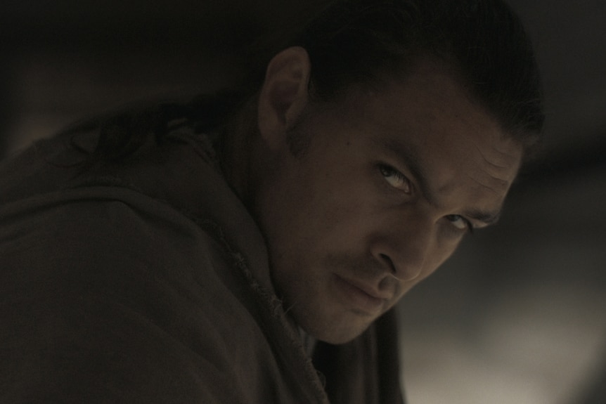 A Native American man in his 40s is being cast in the shadows.  His hair is pushed back and he has an intense look.