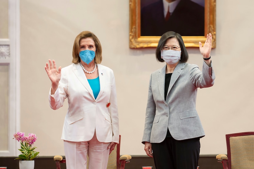 Two older women wearing conservative-looking pantsuits stand and wave to the camera in front of two chairs and a painting.