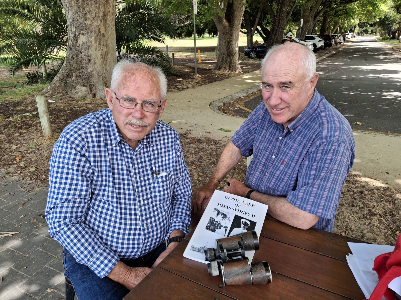 Two older men smile at the camera, a book and binoculars on the table before them.