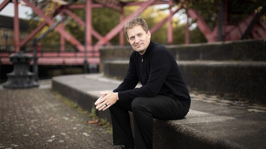 Pianist Andrew Armstrong sits on concrete steps next to a wrough iron brigde wearing a black jumper and trousers.