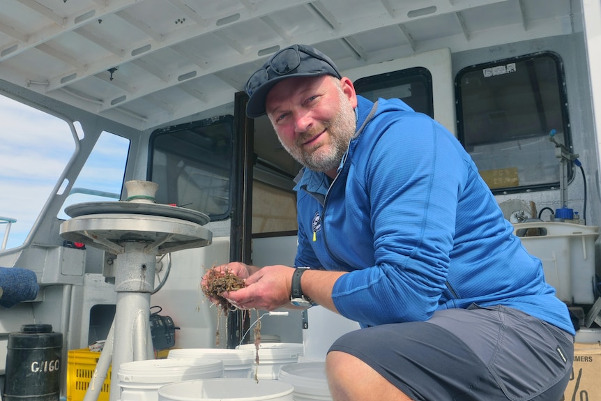 A man, wearing a blue shirt, crouches down on the deck of a boat.