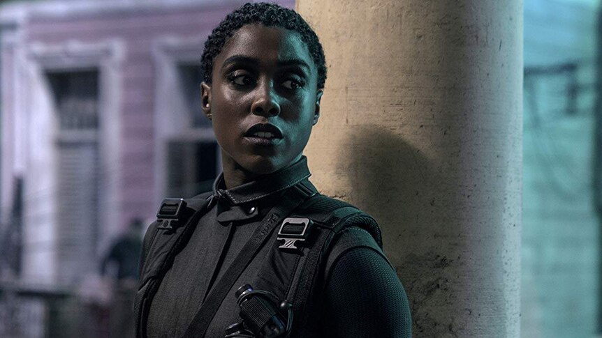 Lashana Lynch looks concerned as she stands near a pole dressed in black, holding a gun