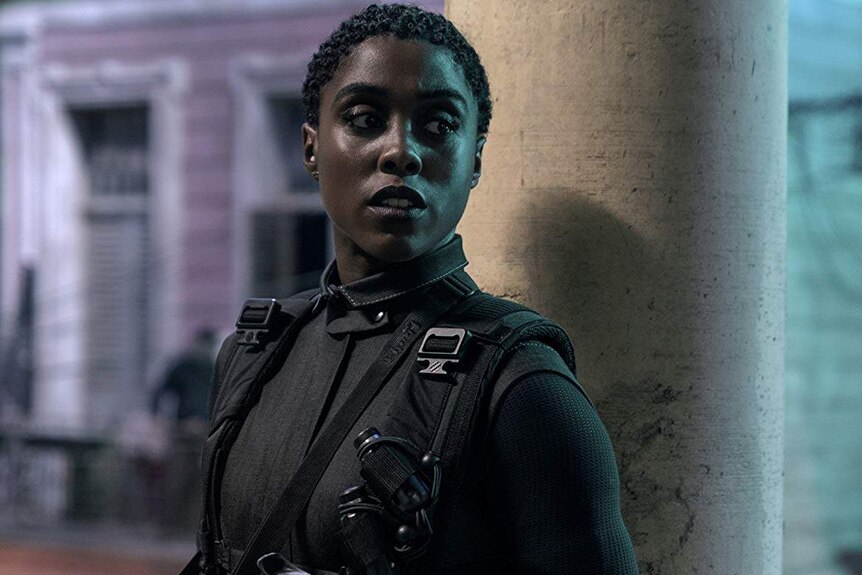 Lashana Lynch looks concerned as she stands near a pole dressed in black, holding a gun