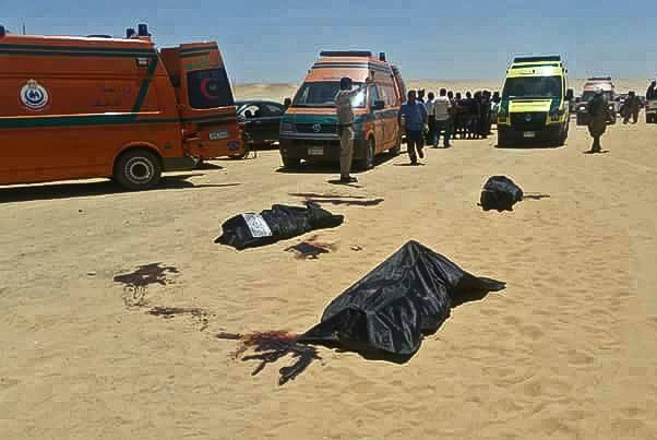 Three bodies lie covered in the desert in front of ambulances and bystanders.