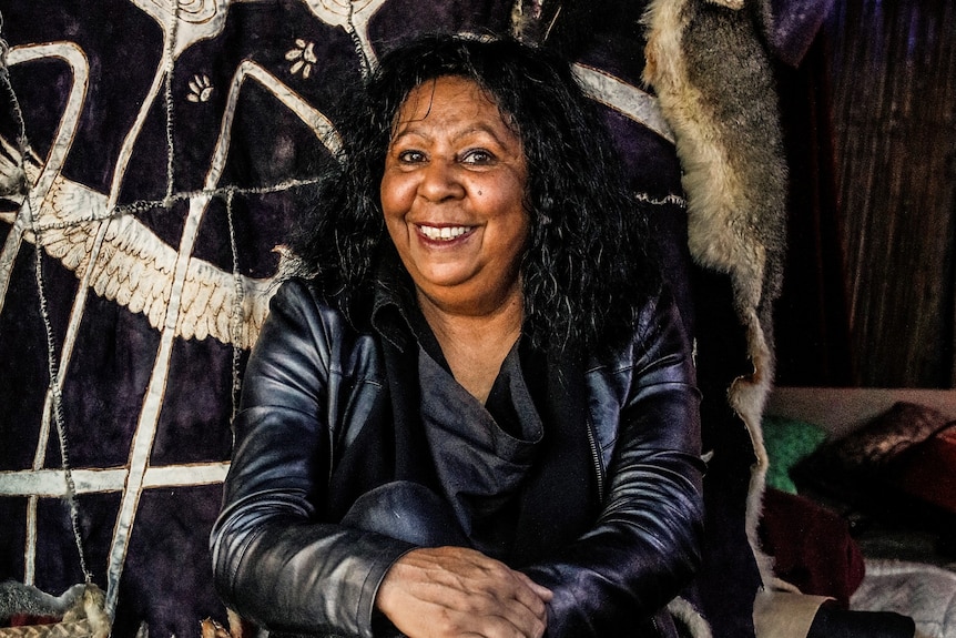 A 60-something Aboriginal woman sits smiling in her studio with an embroidered tapestry behind her