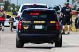 Police vehicles block a road with police tape marking a crime scene in the background