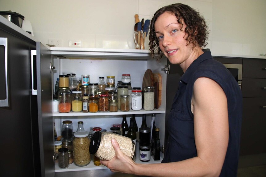 A woman stands in her kitchen next to a full pantry holding a jar of grains and looking at the camera.