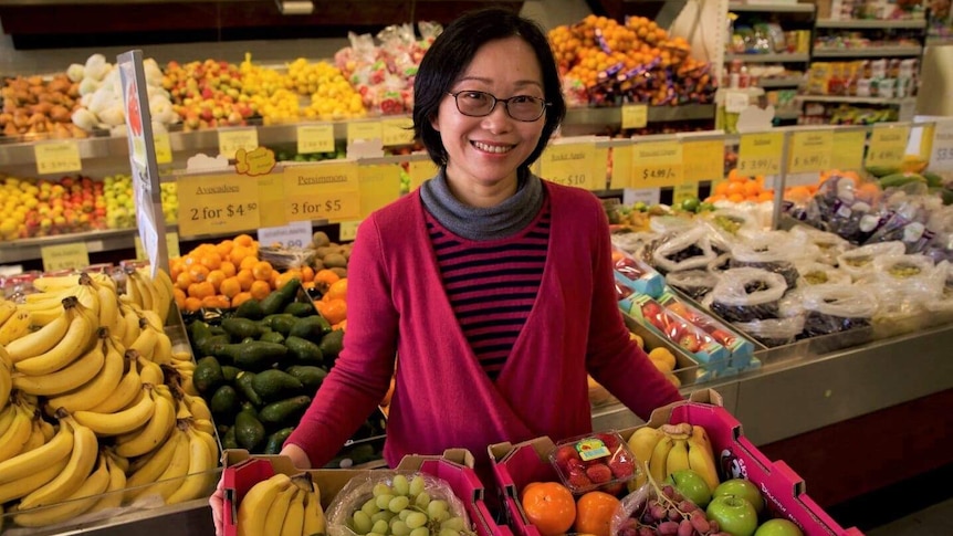 A woman stands holding two boxes full of different fruits. She is in a grocery store.