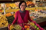 A woman stands holding two boxes full of different fruits. She is in a grocery store.