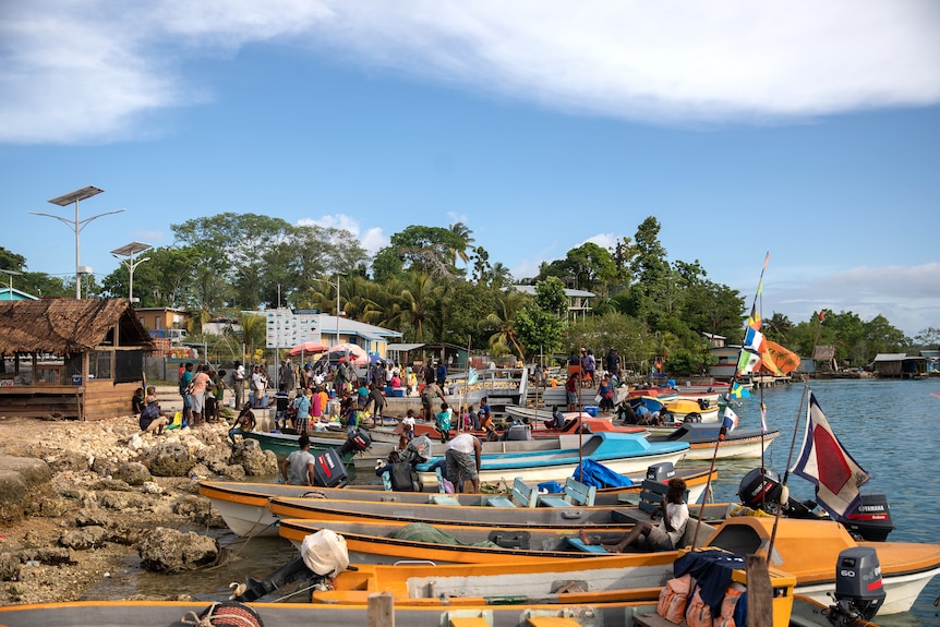A number of small boats on the shore of a rocky beach. Dozens of people are standing and sitting around and on the boats.