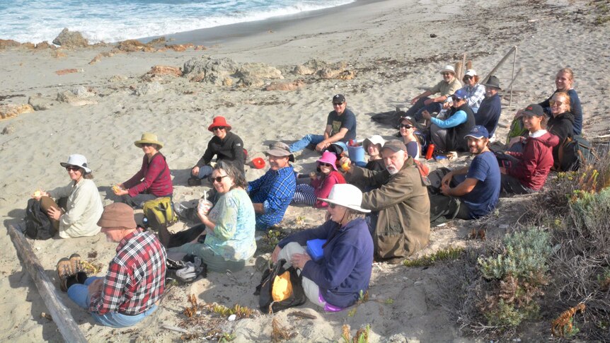 A group of people in foreground sitting on sand with backs to the camera facing an ocean bay.