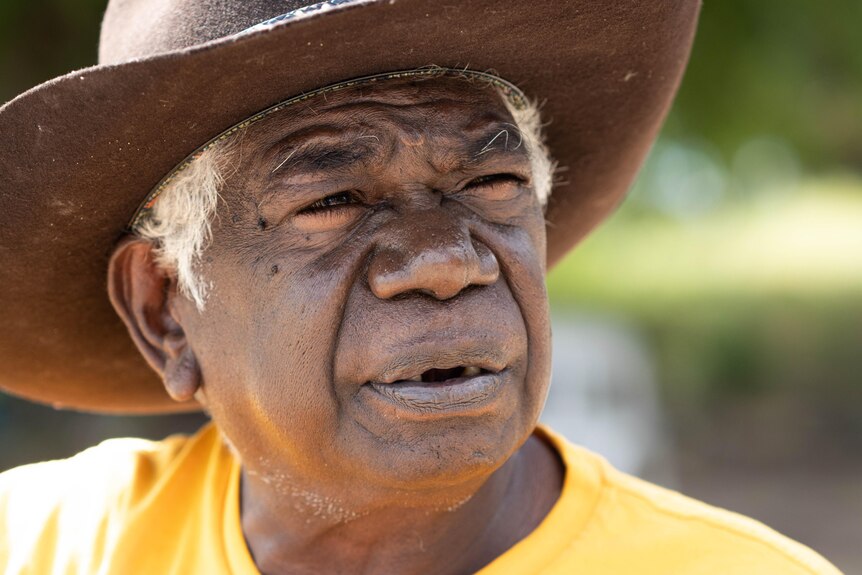 A close-up photo of an Aboriginal man's face. He is wearing a big hat