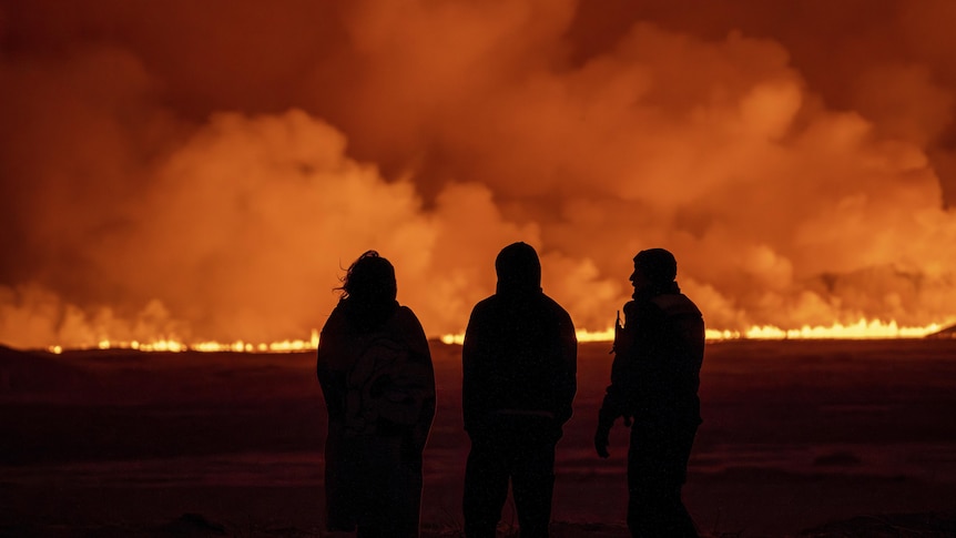 The backs of three people standing in the dark, watching fire and lava spew in a line from a volcanic eruption