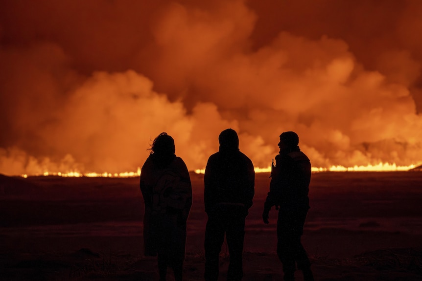 The backs of three people standing in the dark, watching fire and lava spew in a line from a volcanic eruption