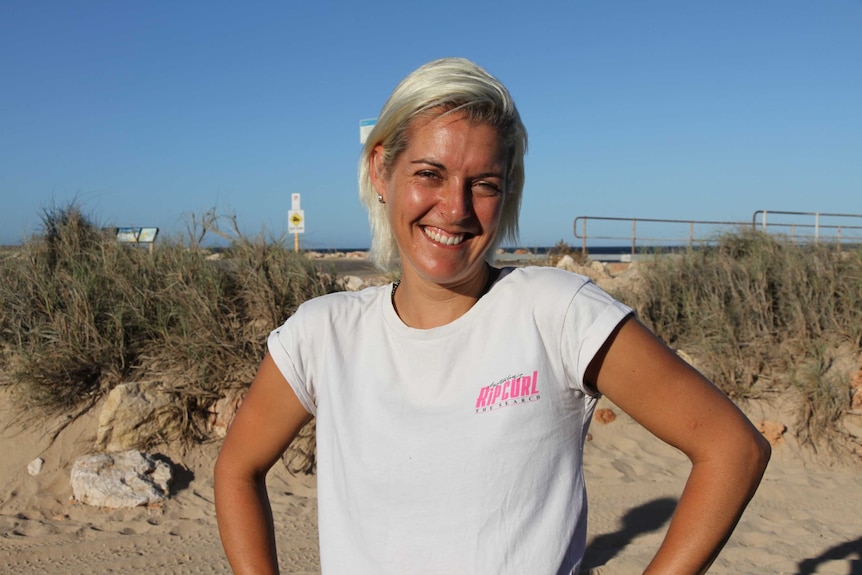 A blonde woman in a white t-shirt grins with her hands on her hips at a beach.