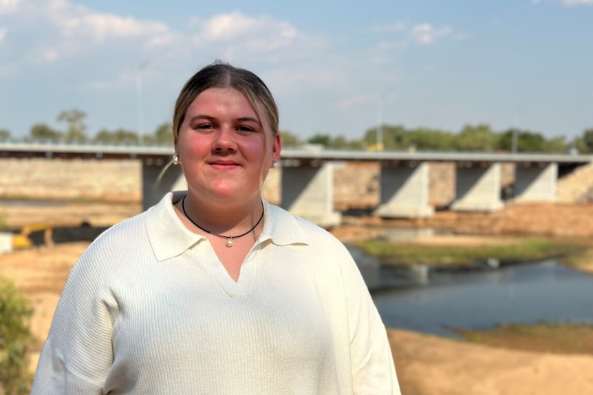 A young woman with white skin and a white t-shirt smiles at the camera with a bridge in the background 