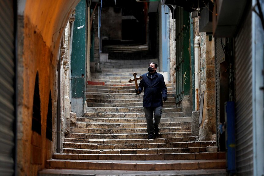 A man wearing a face mask holds a Christian cross as he walks down stone stairs past closed shops.