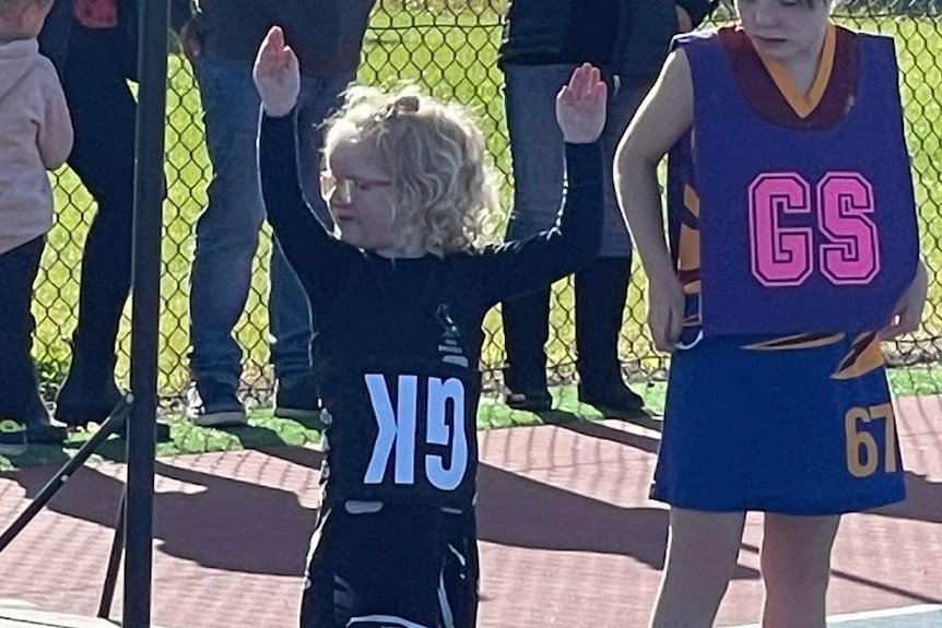 Four-year-old girl with blonde hair and glasses holds her hands above her head on a netball court with an upside down GK bib.