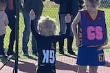 Four-year-old girl with blonde hair and glasses holds her hands above her head on a netball court with an upside down GK bib.